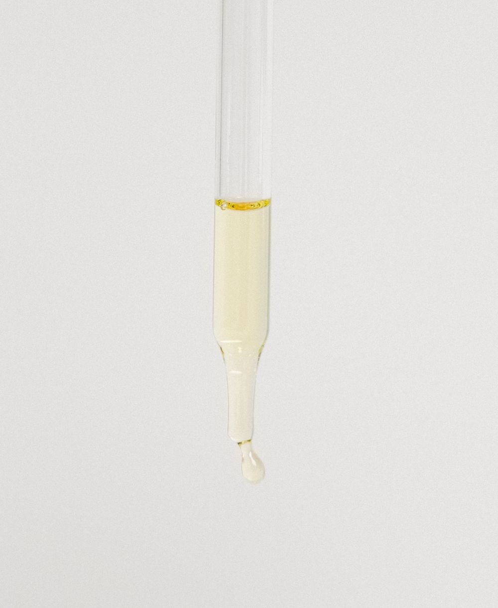 Close-up of a glass dropper pipette dispensing a drop of clear serum on a white background.