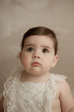 Baby wearing delicate white lace dress looking up with wide-eyed expression against neutral background.