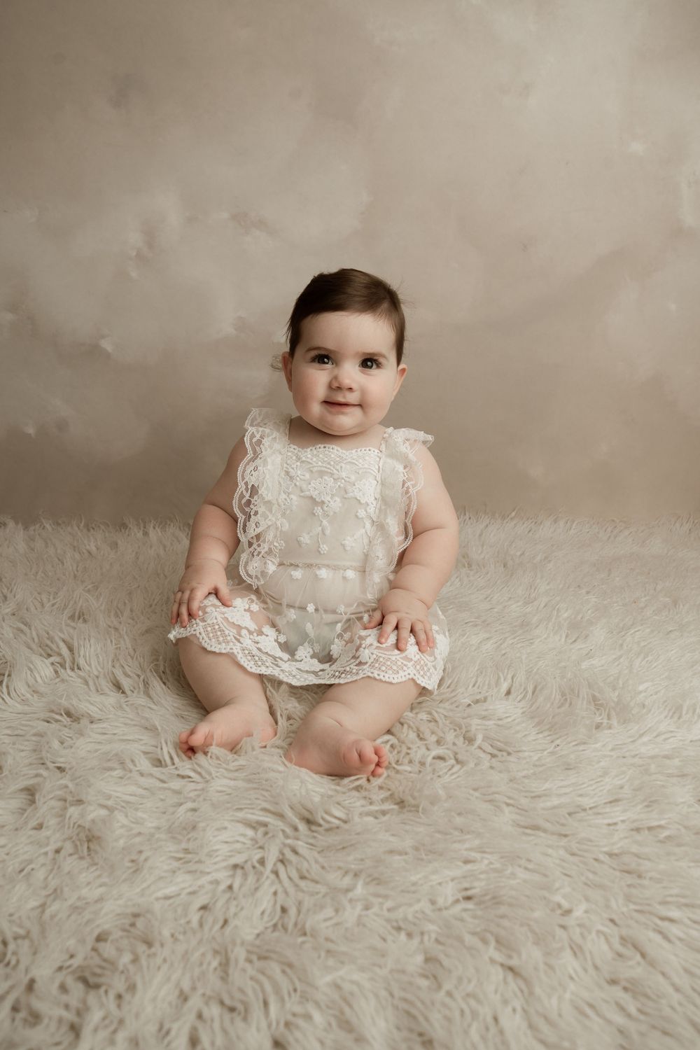 A baby wearing a white lace dress sits on a fluffy white rug in a professional studio portrait setting.