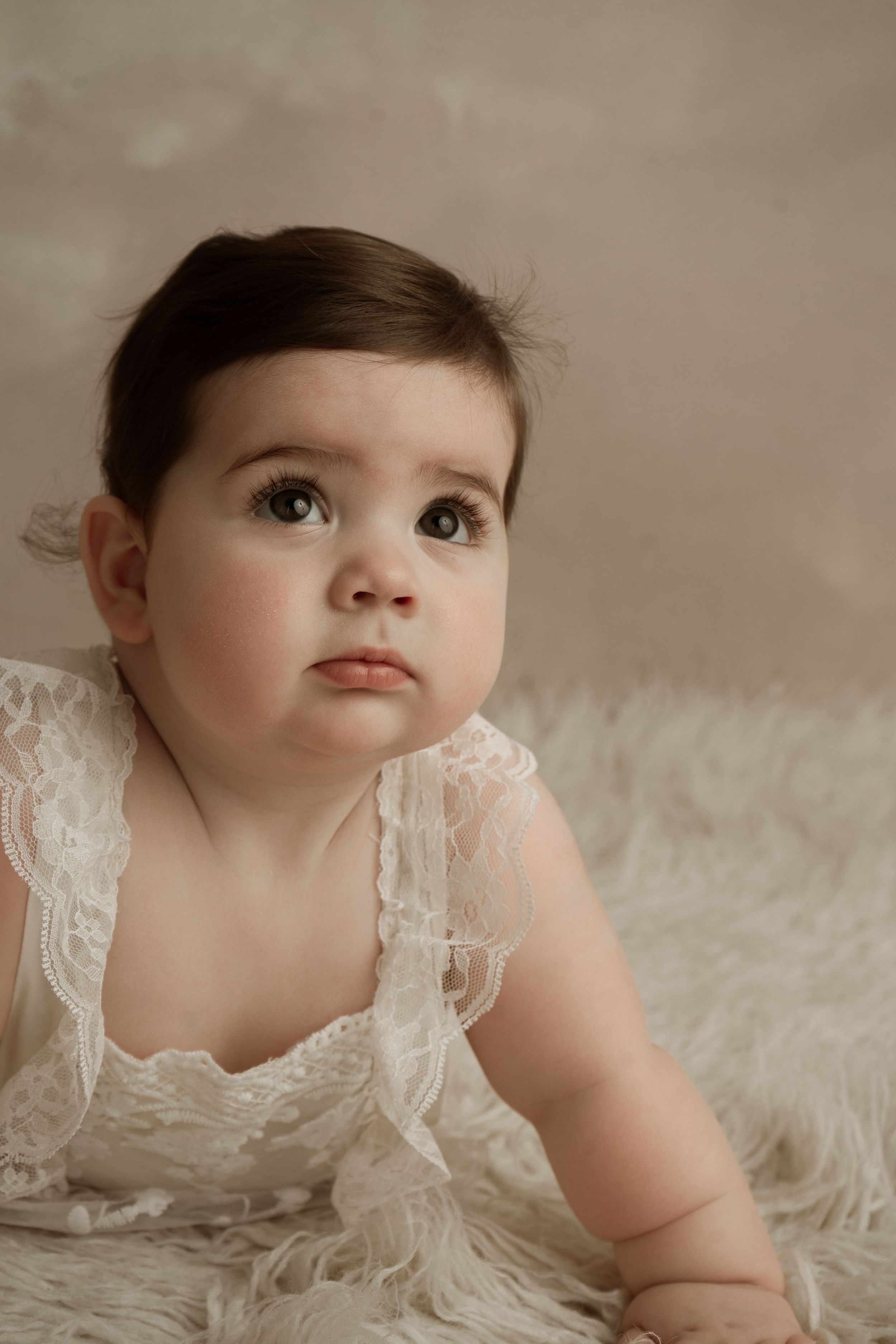 Baby in white lace dress laying on soft white fur blanket in artistic portrait series.