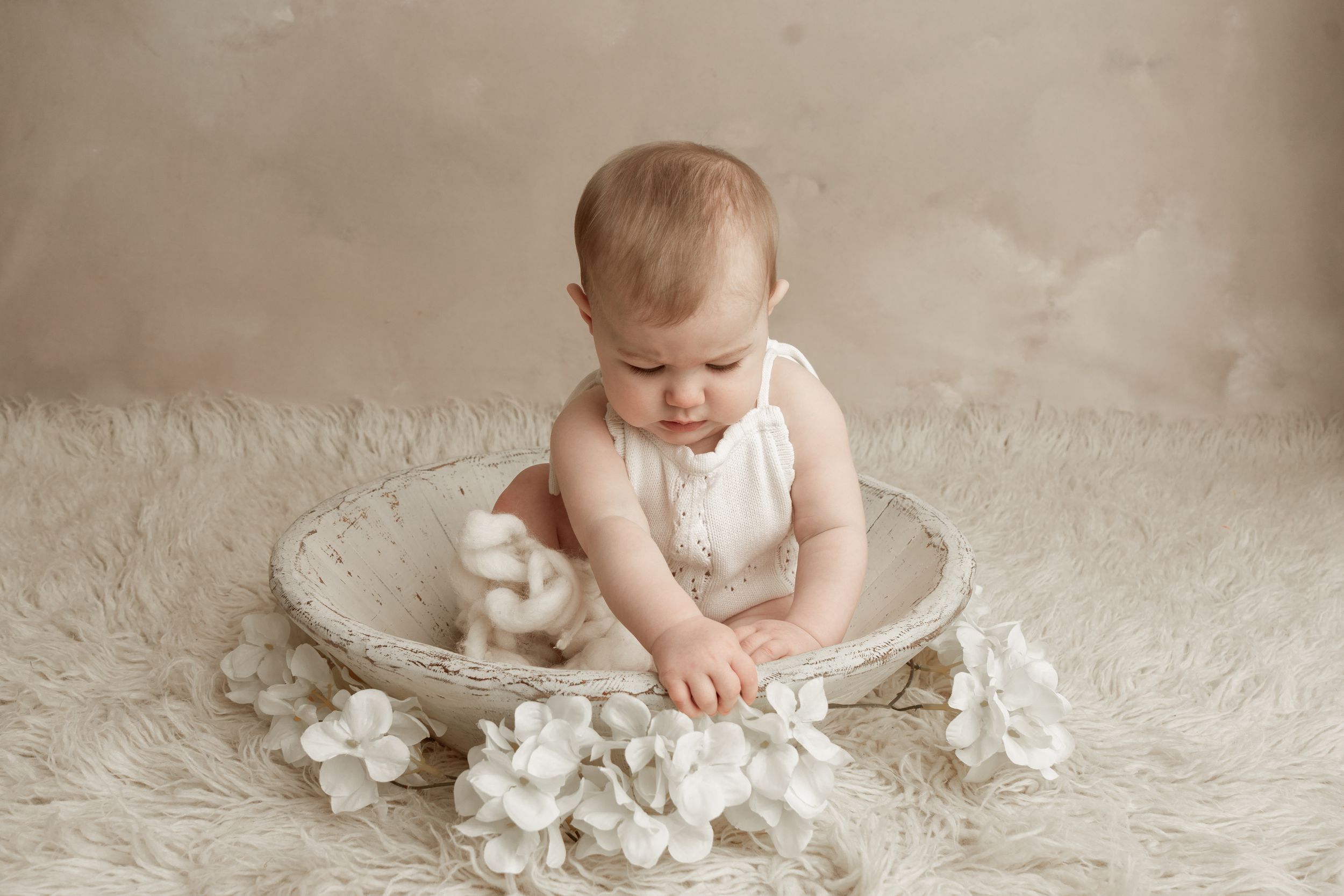 Baby wearing white sits on plush carpet surrounded by soft tissue paper in a light-filled studio setting.