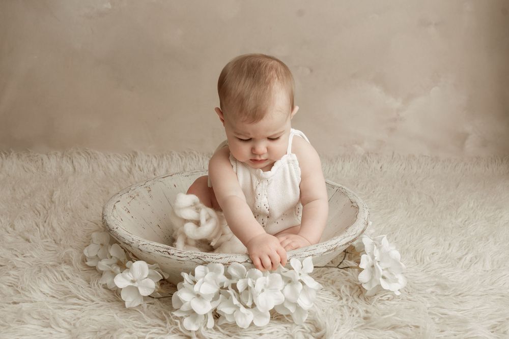 Baby wearing white sits on plush carpet surrounded by soft tissue paper in a light-filled studio setting.