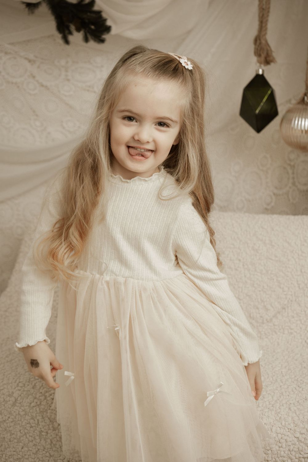 A young child in a cream-colored long sleeve dress stands in front of a white backdrop with holiday decorations.