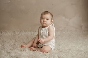 Baby in white sleeveless outfit sits on soft fluffy white rug against neutral beige backdrop during portrait session.