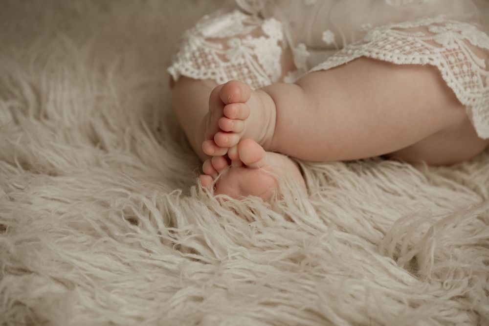 Tiny baby feet resting on soft white fur blanket with lace dress detail visible.