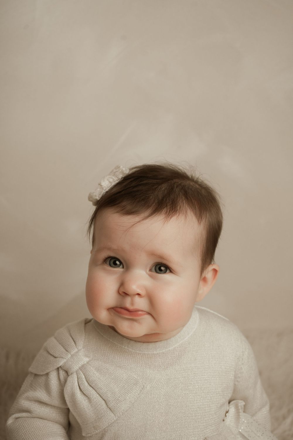 A portrait of an adorable baby wearing a white sweater against a neutral beige background.
