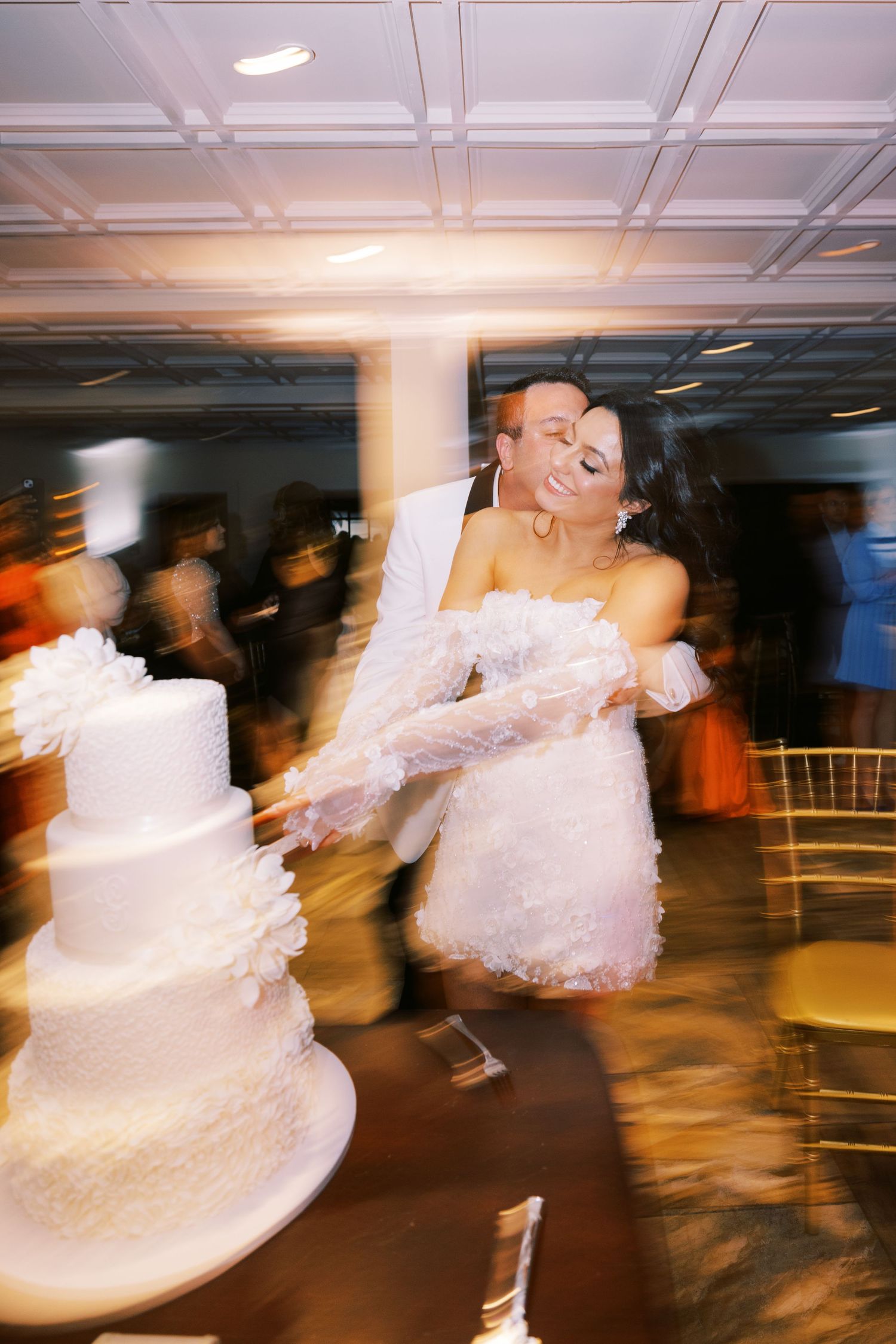 Bride and groom cut elegant white tiered wedding cake during blurred motion reception photo.