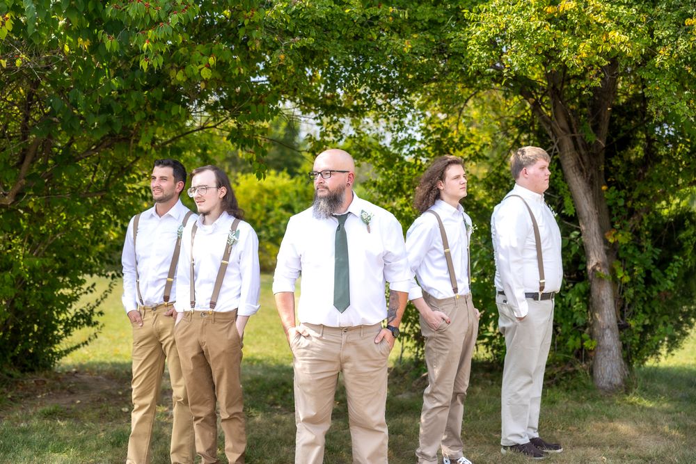 Wedding party members in khaki pants and white shirts with suspenders pose together in a lush outdoor garden setting.