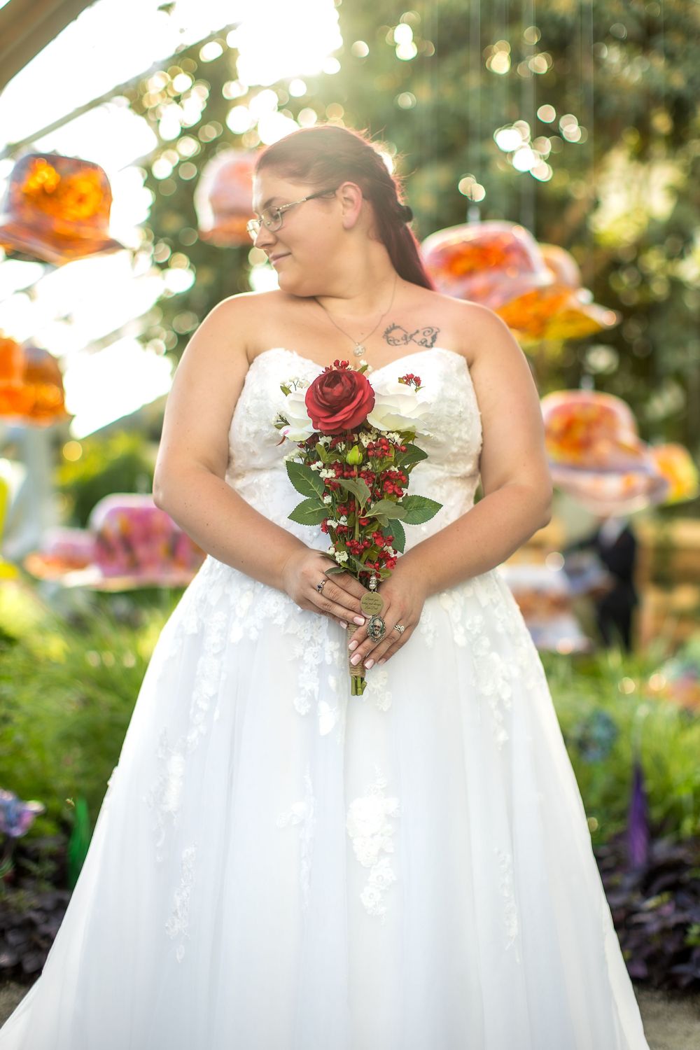 Bride in white wedding gown holding red roses in garden setting with orange decorations overhead.