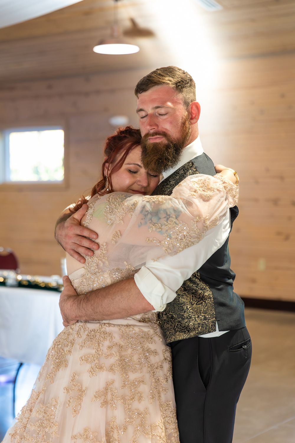 A couple shares an intimate embrace during their first dance at a rustic indoor wedding venue.