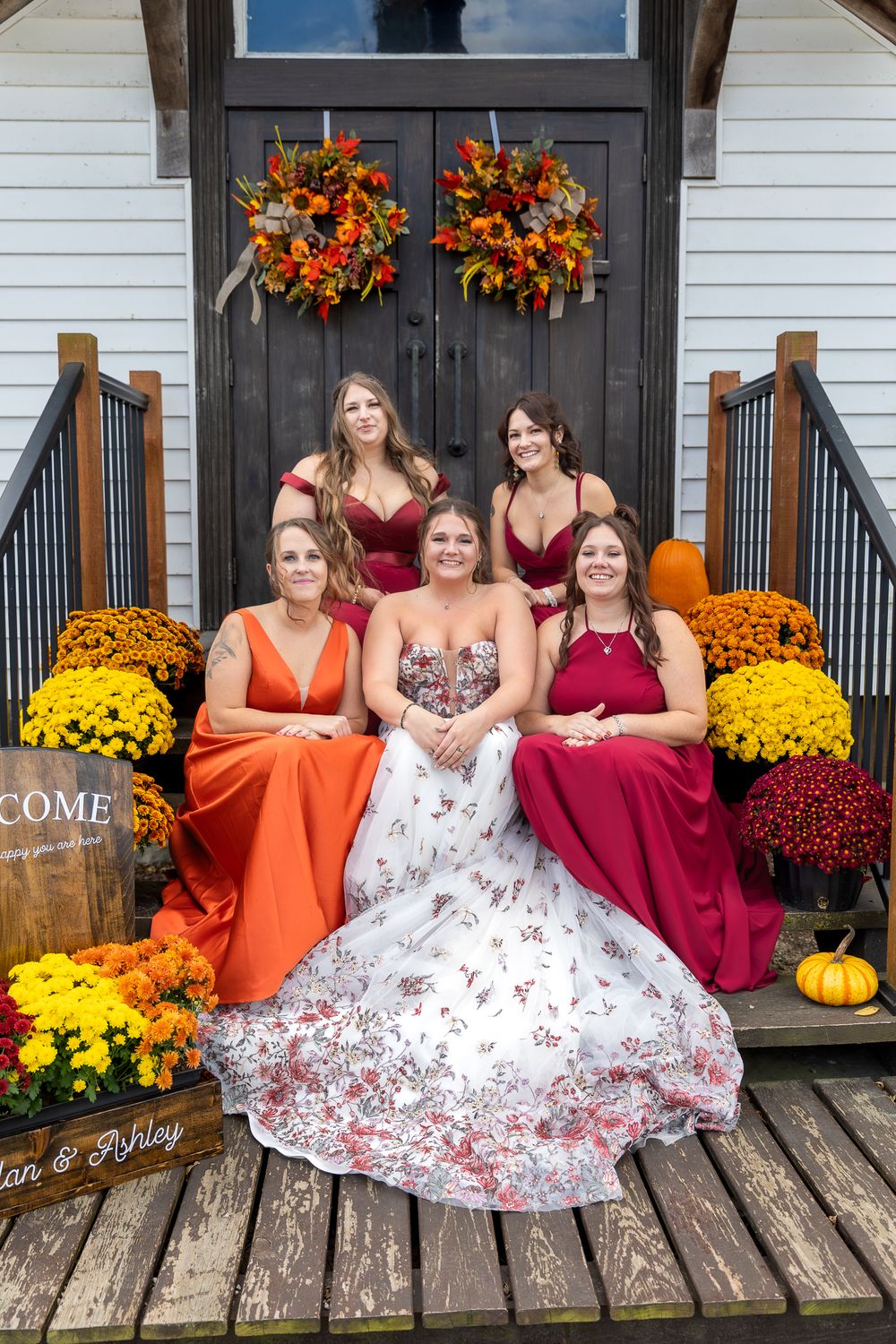 A group in orange and burgundy dresses pose on rustic wooden steps decorated with yellow mums and fall decor.