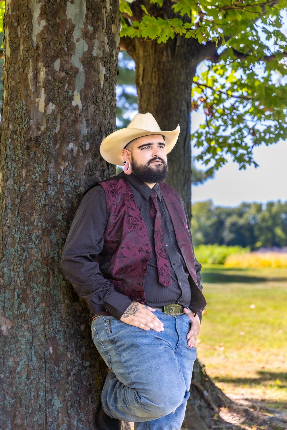 A person in western attire and cowboy hat leans against a large tree trunk in a sunny park.