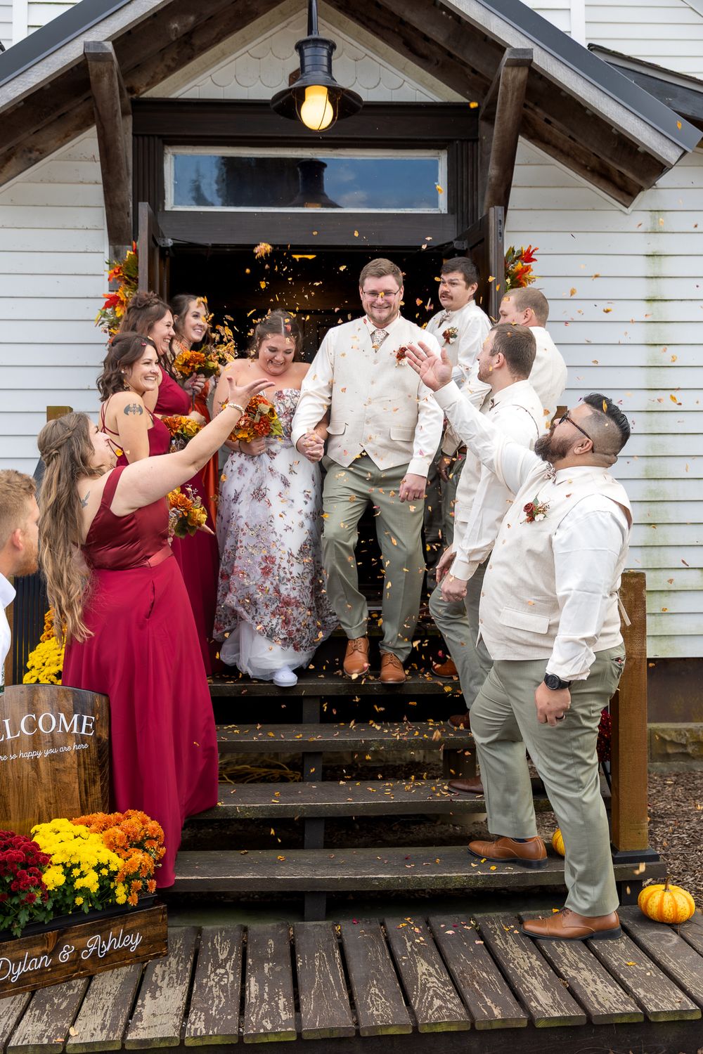 Wedding party celebrates with champagne spraying on rustic wooden porch decorated with autumn flowers.