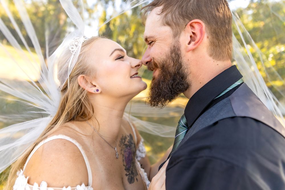 A couple in wedding attire share a romantic moment outdoors in golden sunlight under a flowing veil.