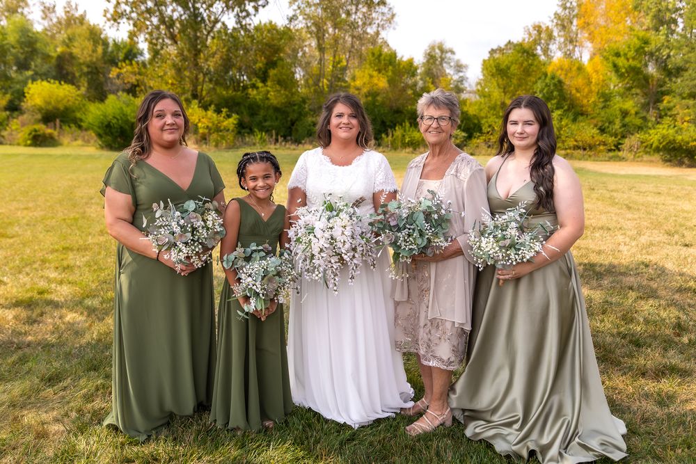 A group stands together in a field during autumn holding sage green and white floral bouquets.