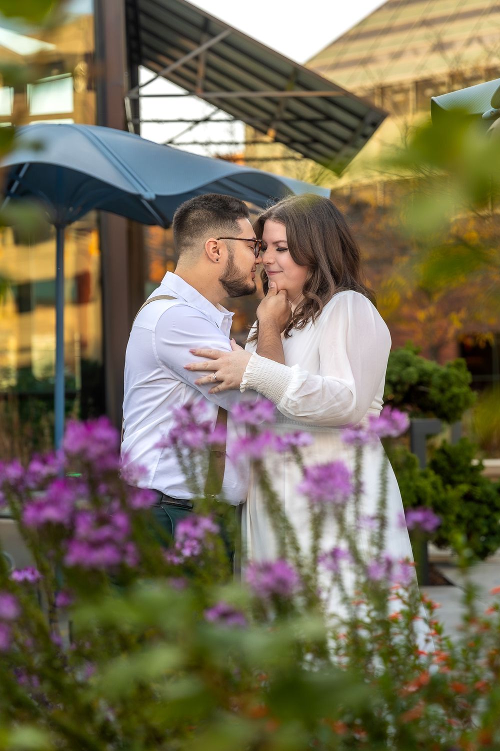 Tender embrace between couple in garden with purple flowers and umbrella in background.