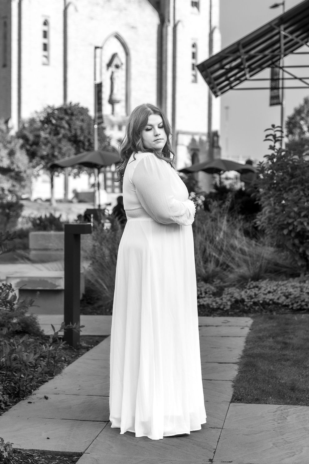 Black and white portrait of person in flowing white dress standing near church architecture.