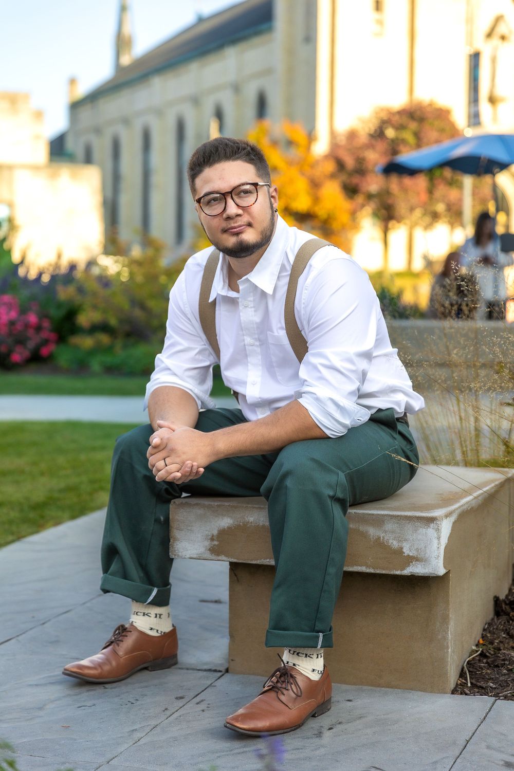 A person in business casual attire with suspenders sits on stone steps near autumn foliage.