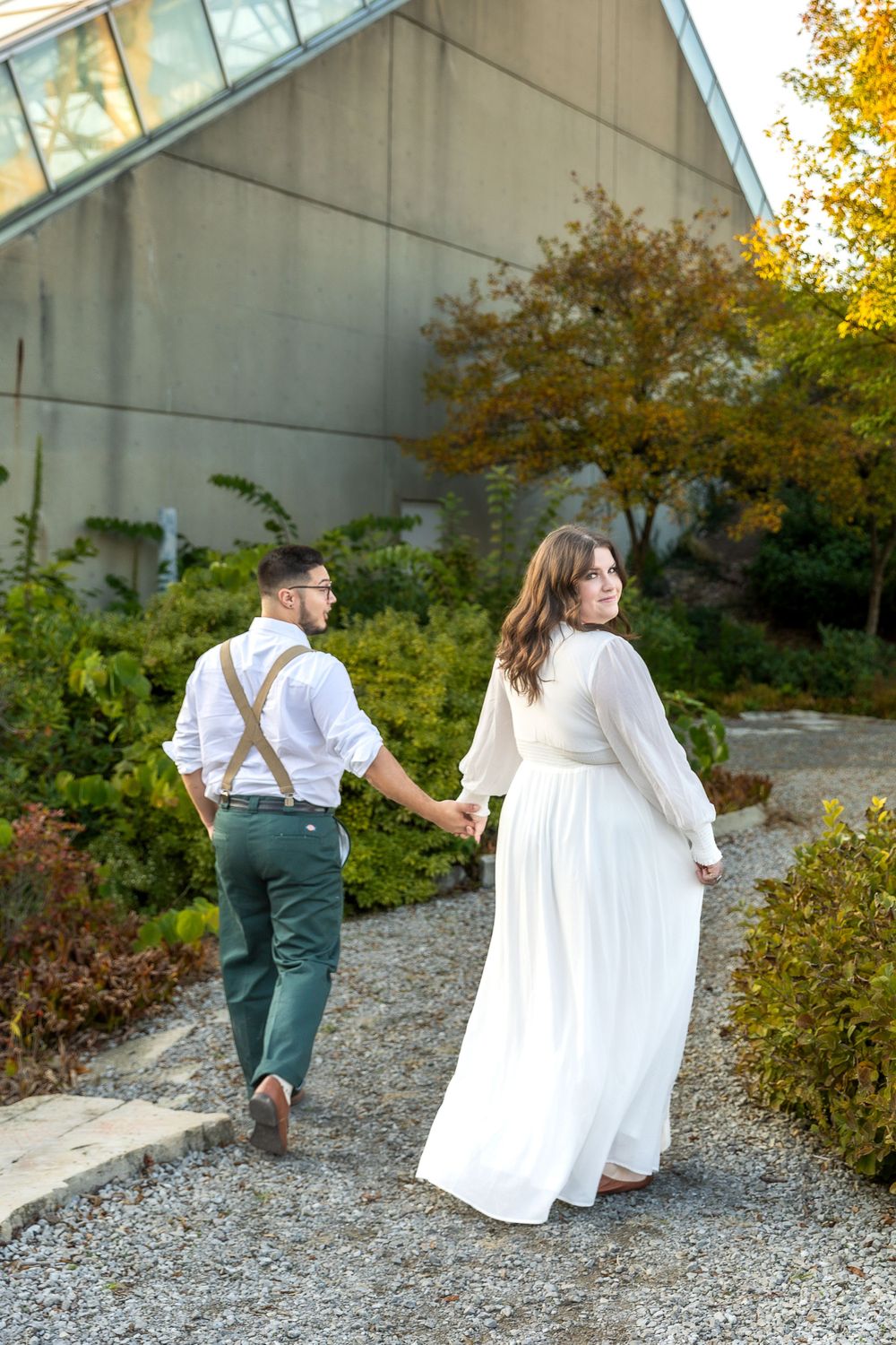 A couple walks hand in hand through a garden path, wearing formal attire with autumn colors in background.