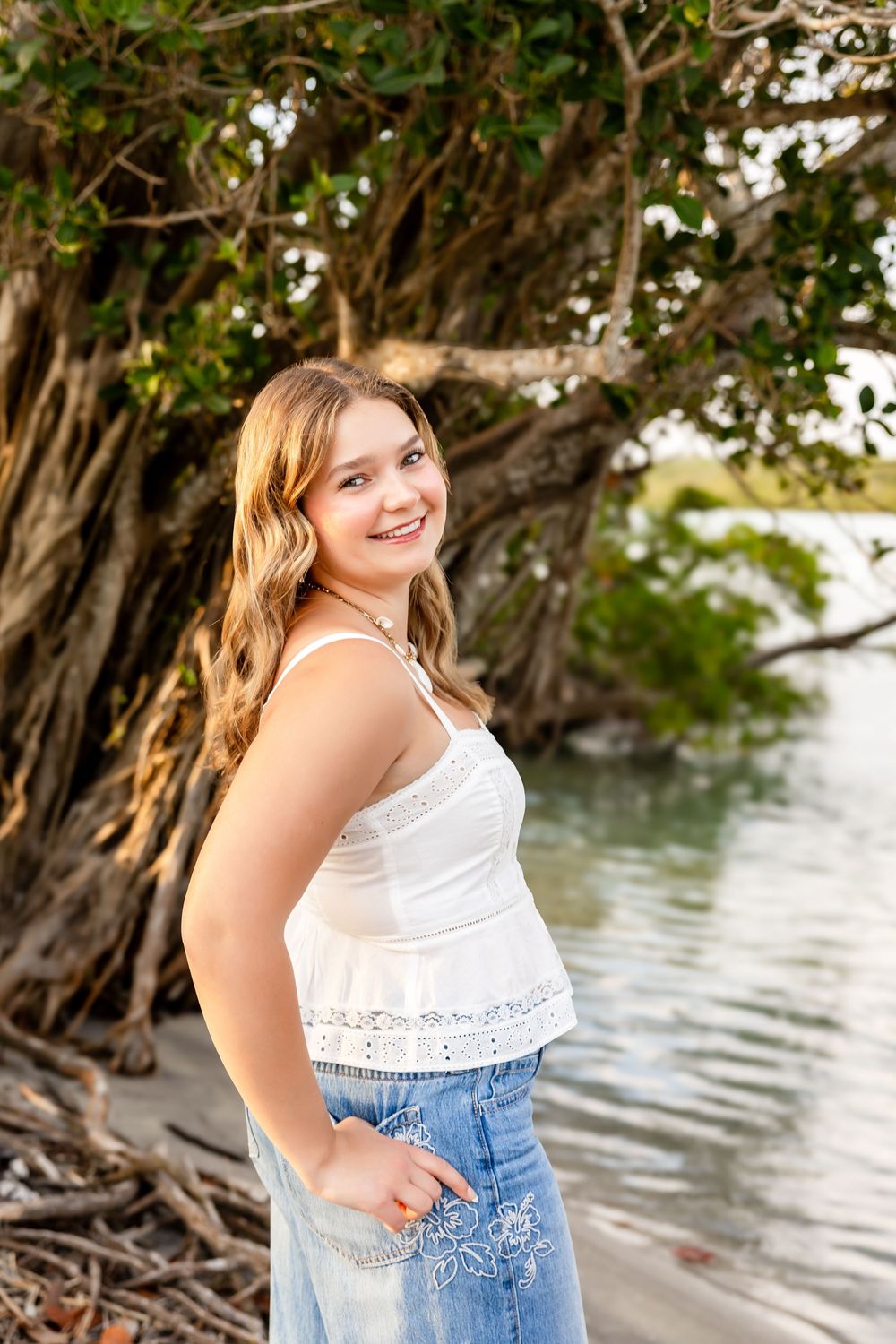 Smiling teenage girl in white top and floral jeans posing by mangrove trees near the water.