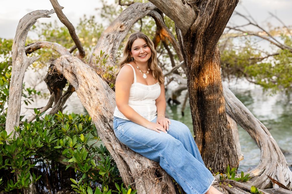 Smiling young woman in white top and jeans sitting on driftwood roots by the water at golden hour.