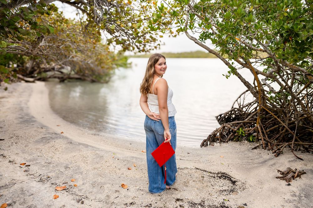 Smiling young woman in jeans holding a red clutch, posing on a sandy shore beneath mangrove trees.