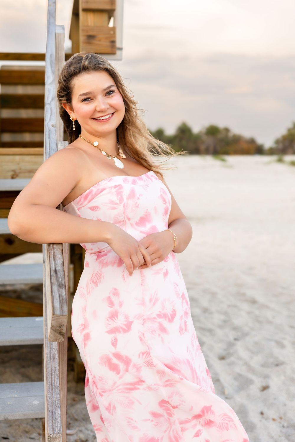 Smiling young woman in a strapless pink floral maxi dress leaning against wooden stairs on a beach.