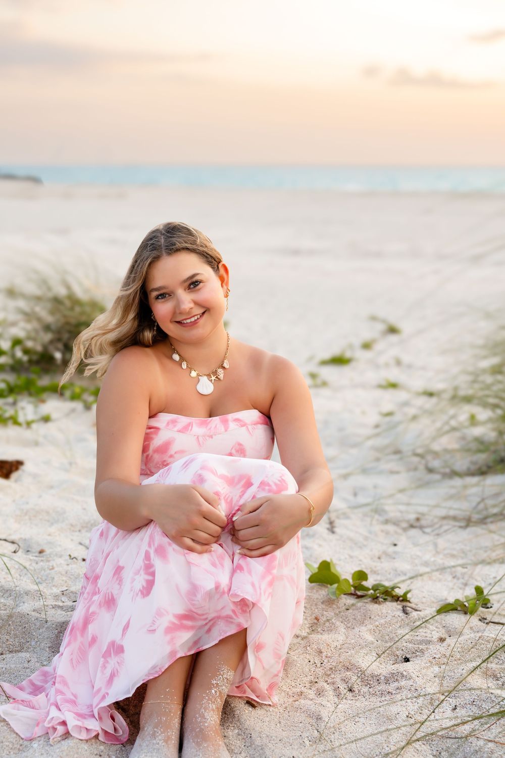 Smiling young woman in a pink floral strapless dress sitting on white sand at a beach sunset.