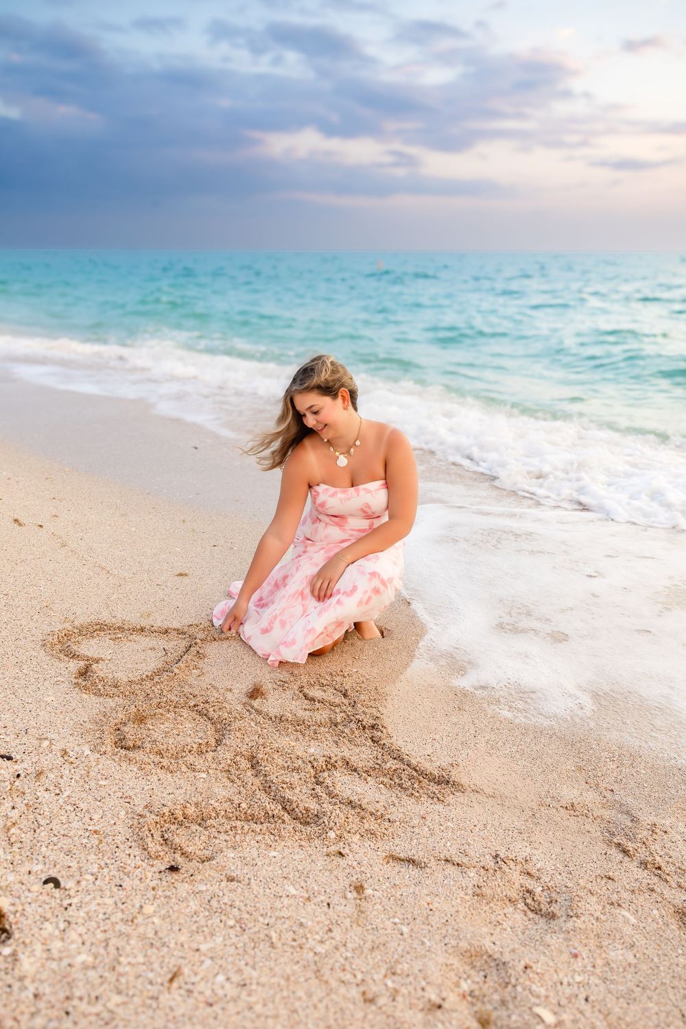 Young woman in a pink floral dress writing in the sand at the ocean's edge during sunset.