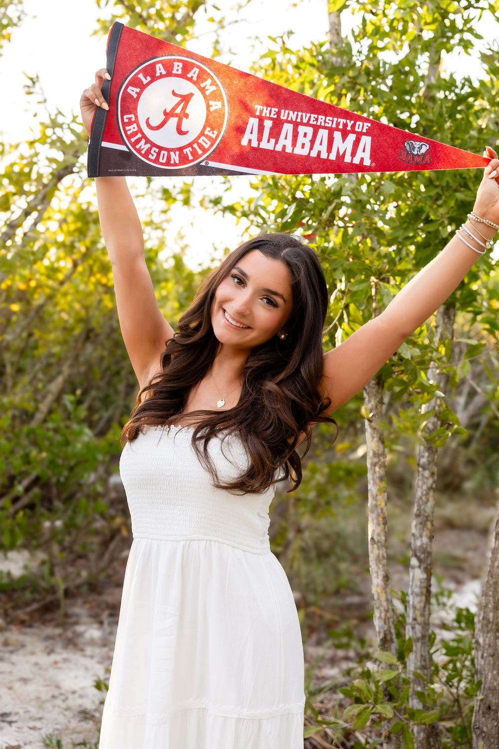 Smiling young woman in white dress holding University of Alabama pennant flag outdoors.