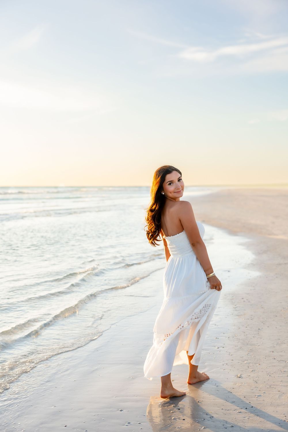 Young woman in white strapless dress walking along ocean shoreline at golden sunset.