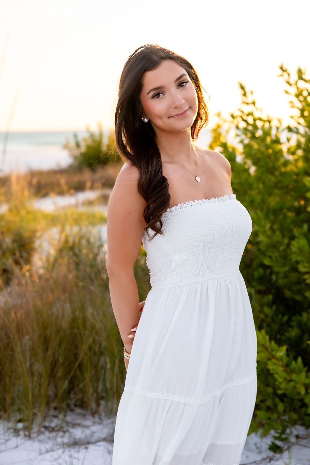 Beautiful young woman in white strapless dress posing among beach dunes at sunset.