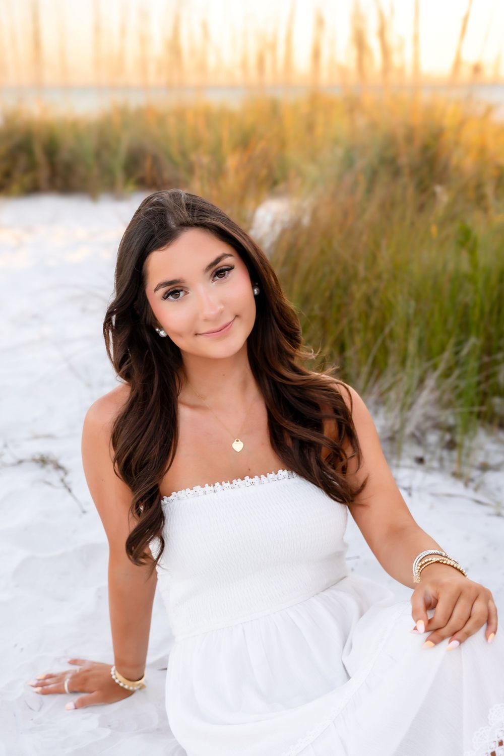 Young woman with long dark hair in white dress sitting on white sand dunes at sunset.