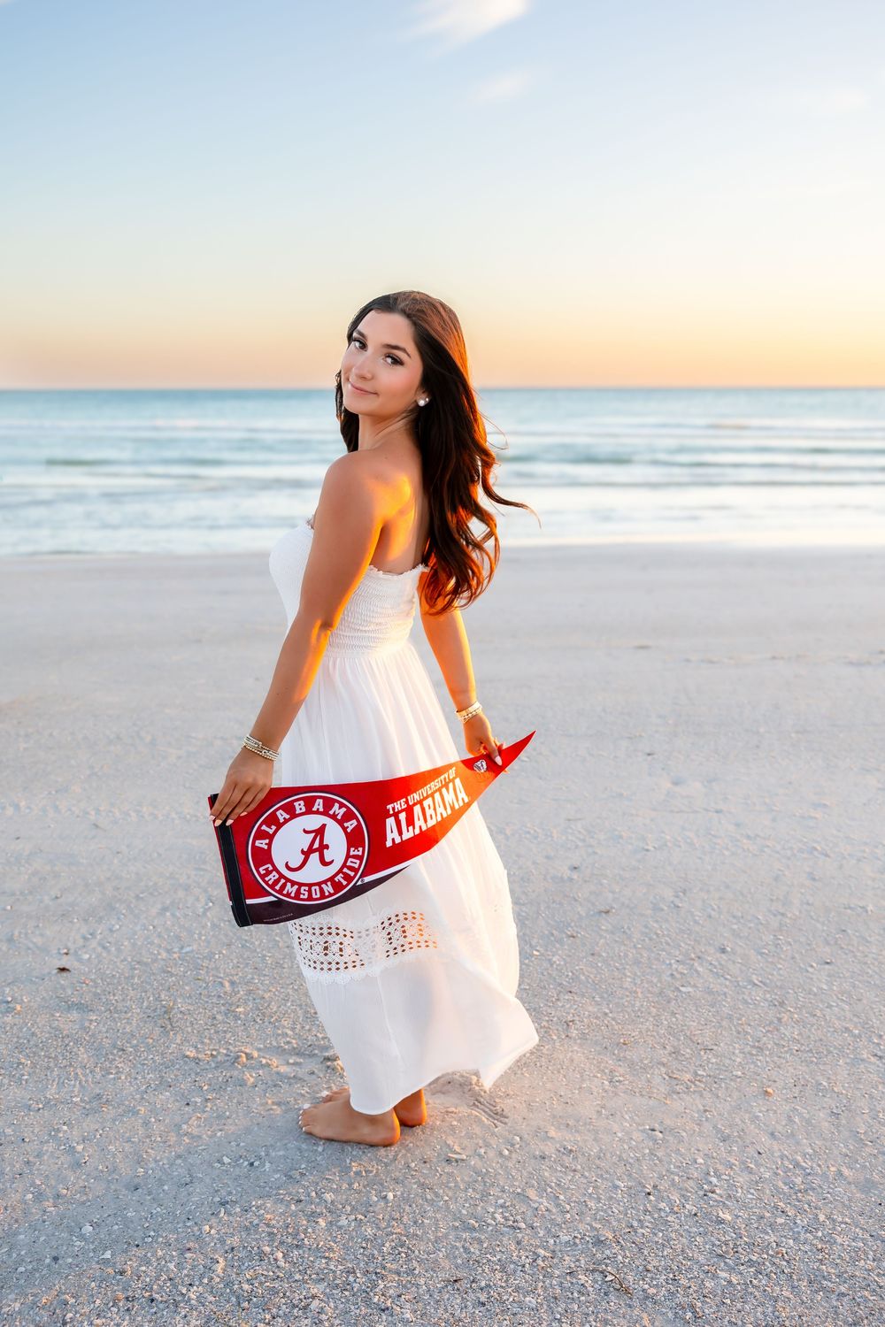 Young woman in white dress holding University of Alabama pennant on beach at sunset.