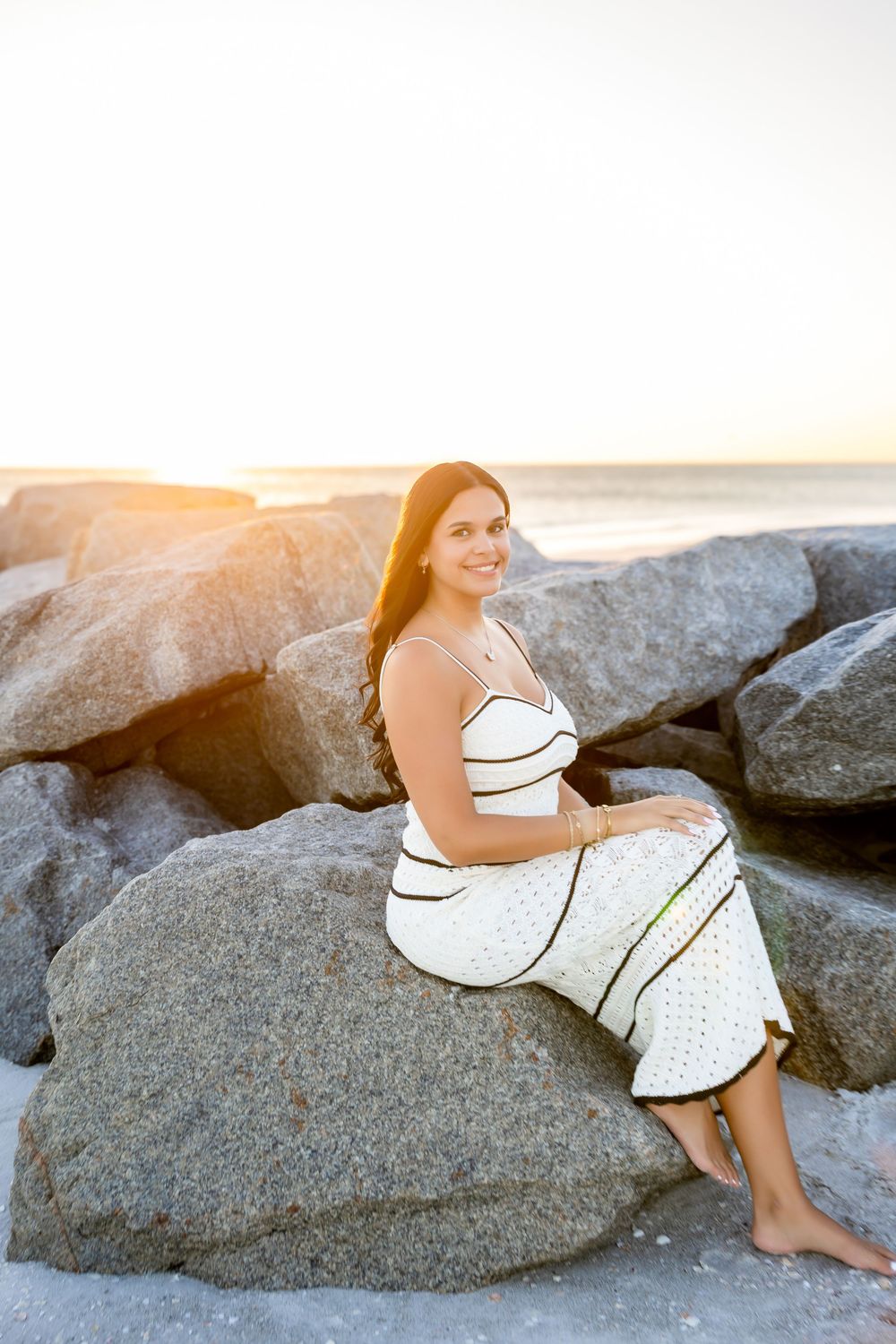 Smiling young woman in white striped dress sitting on large rocks at beach during golden hour sunset.