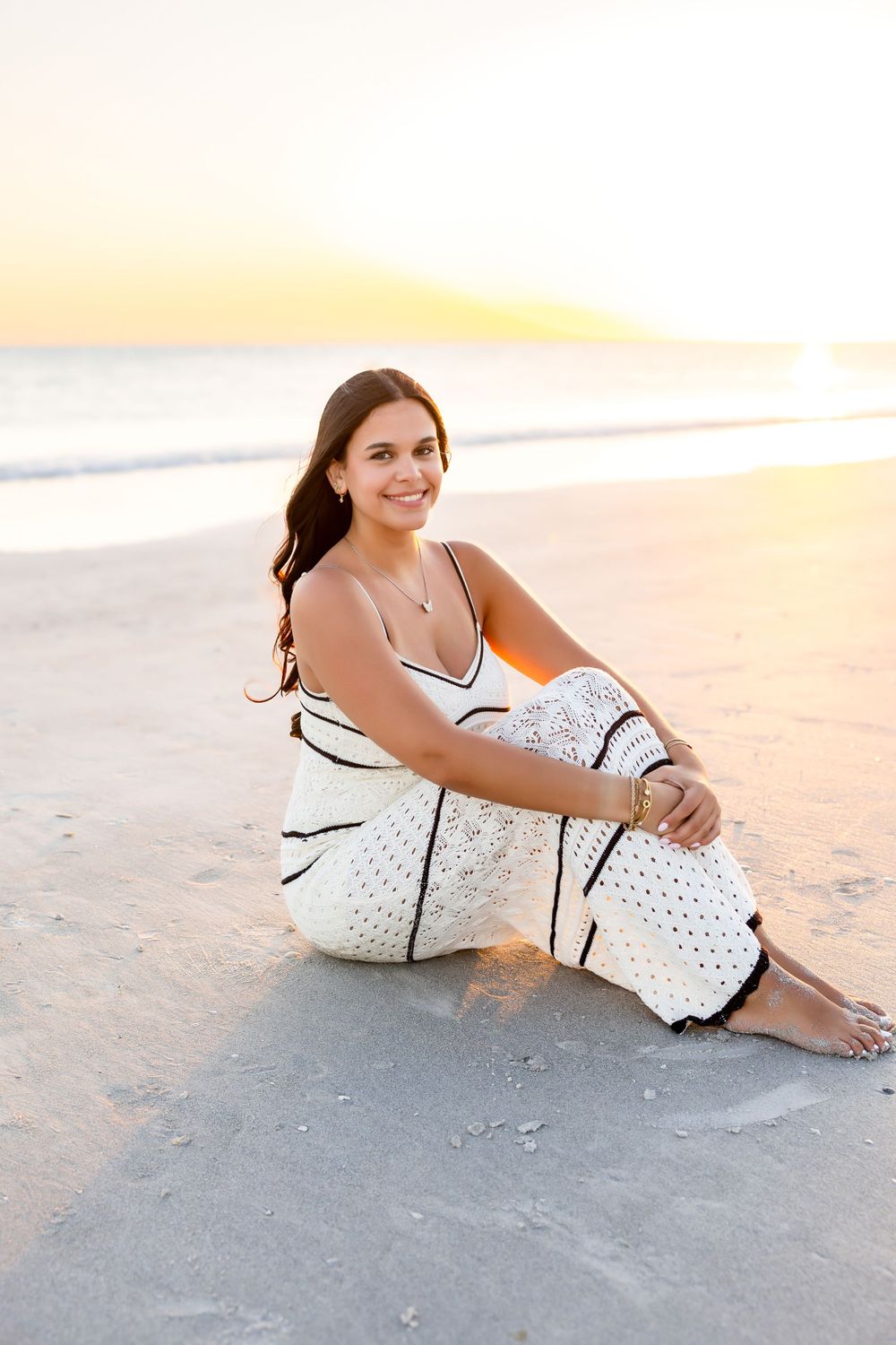 Young brunette woman in white patterned jumpsuit sitting on beach sand at golden sunset by the ocean.