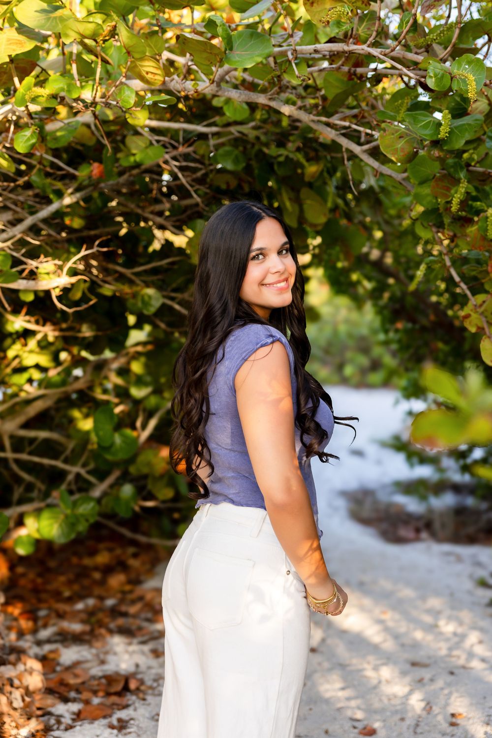 Young woman with long dark hair in blue top and white pants smiling on a sandy beach path under green trees.