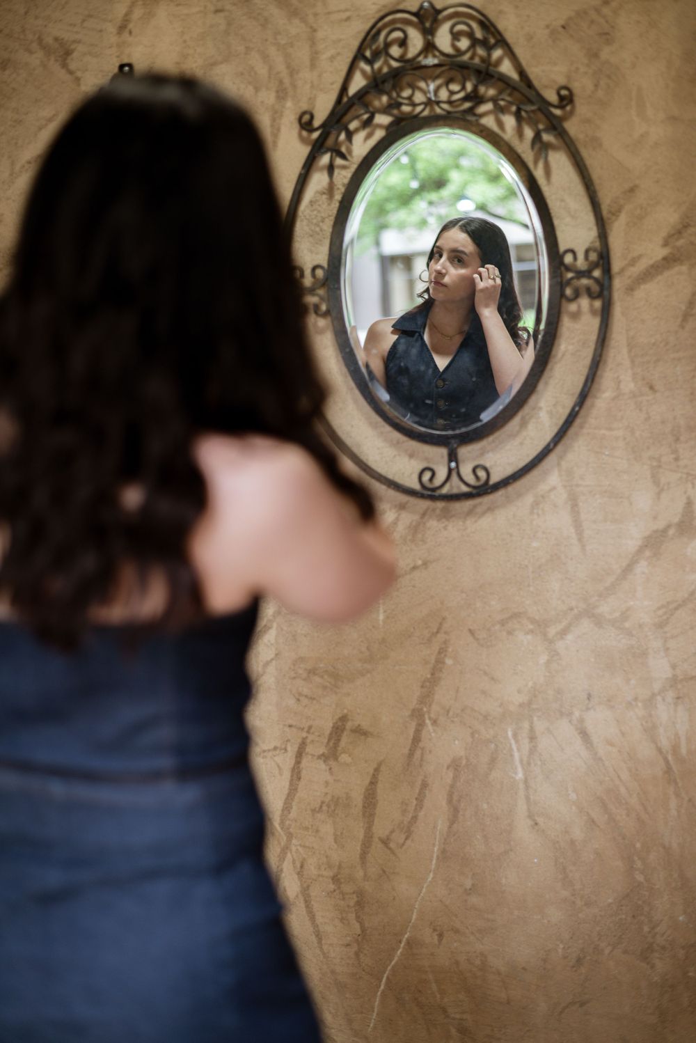 An artistic mirror reflection shot showing a woman adjusting her hair against a textured wall.