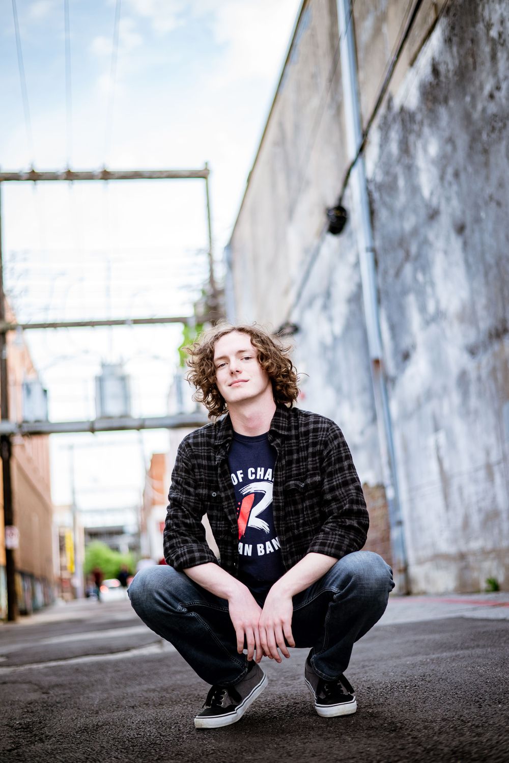 Person crouching in urban alley wearing plaid shirt and graphic tee.