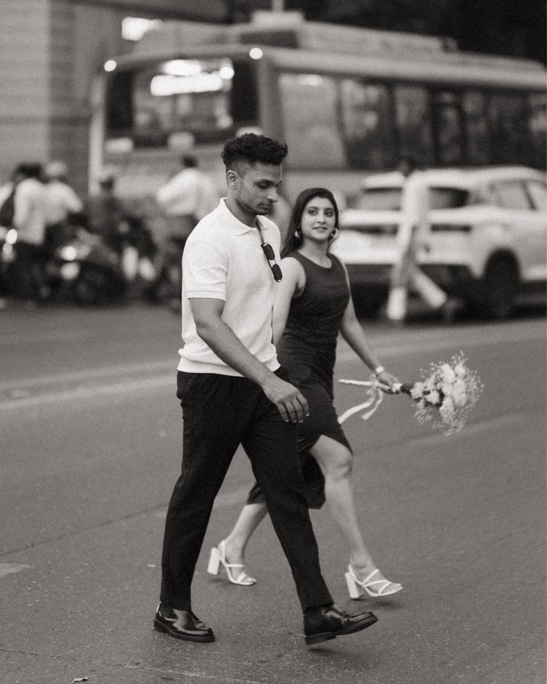 A romantic black and white photo of a couple walking on a street, with one holding a bouquet of flowers.