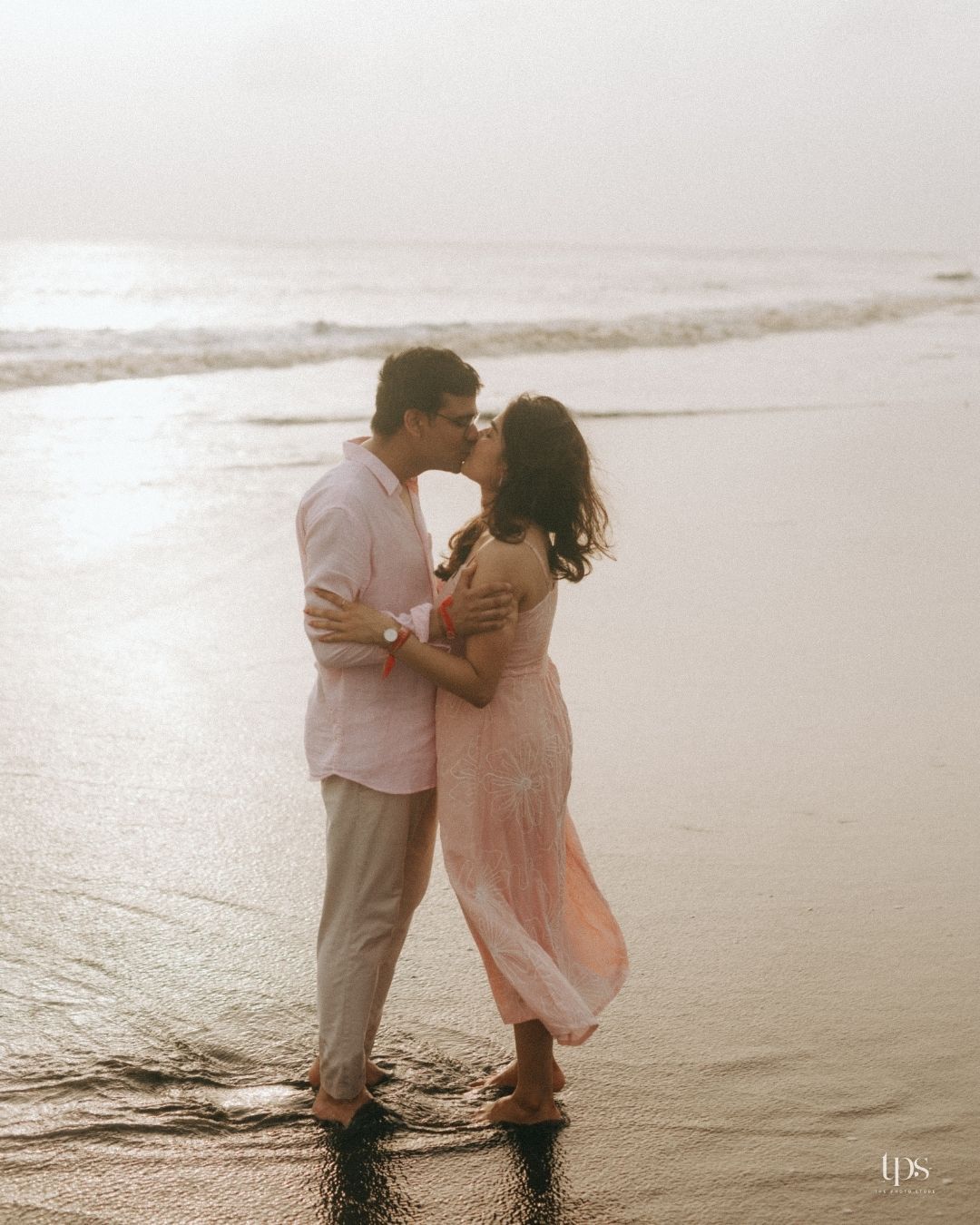 Couple in light pink and white outfits sharing romantic moments on a misty beach.