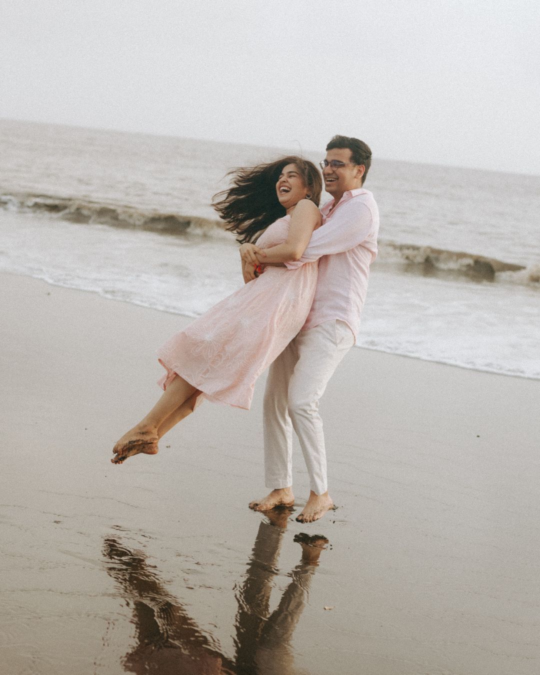 Playful beach photoshoot with couple in pink dress and white outfit splashing in waves.