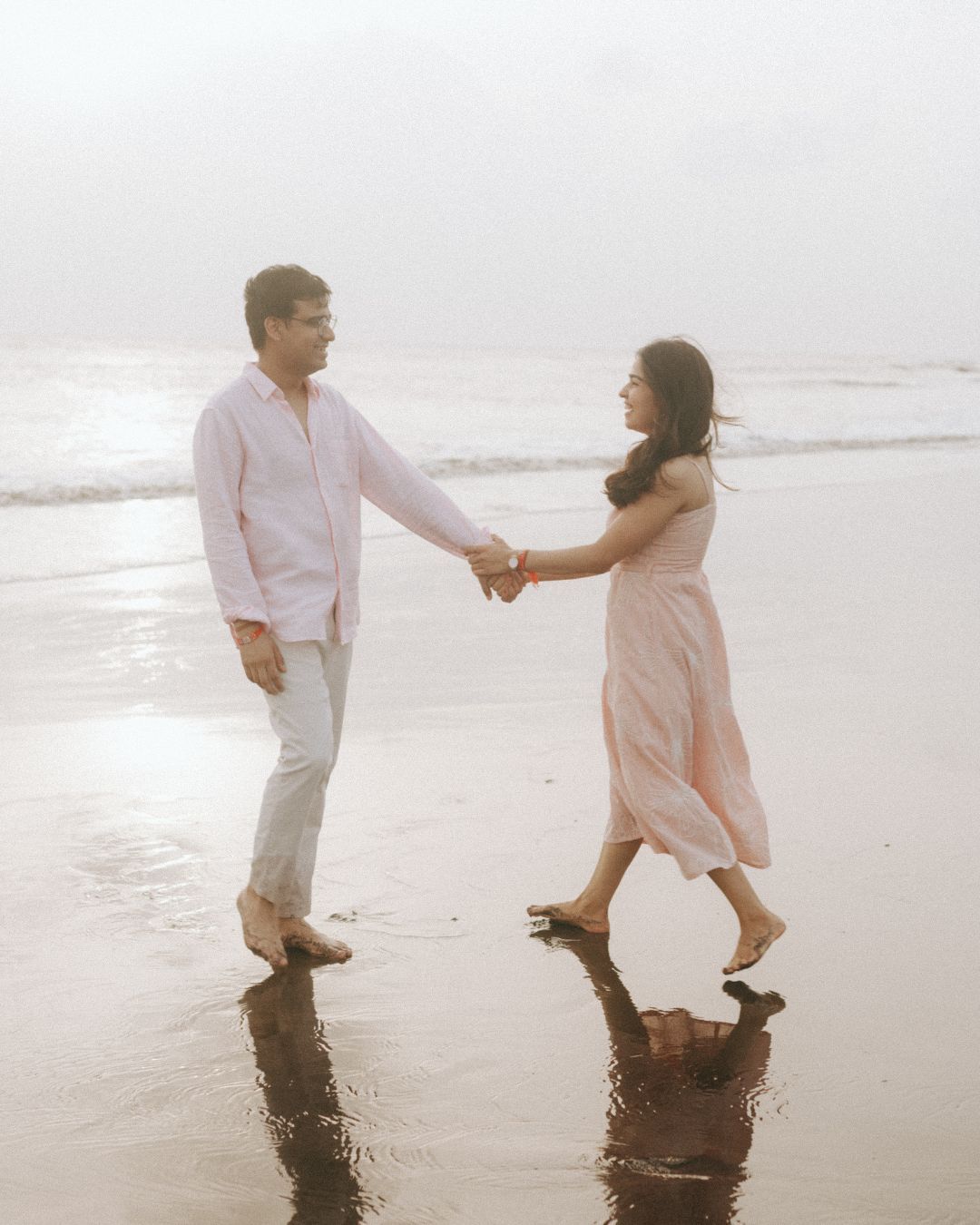 Couple walking and dancing together on beach shore in pink and white casual outfits.
