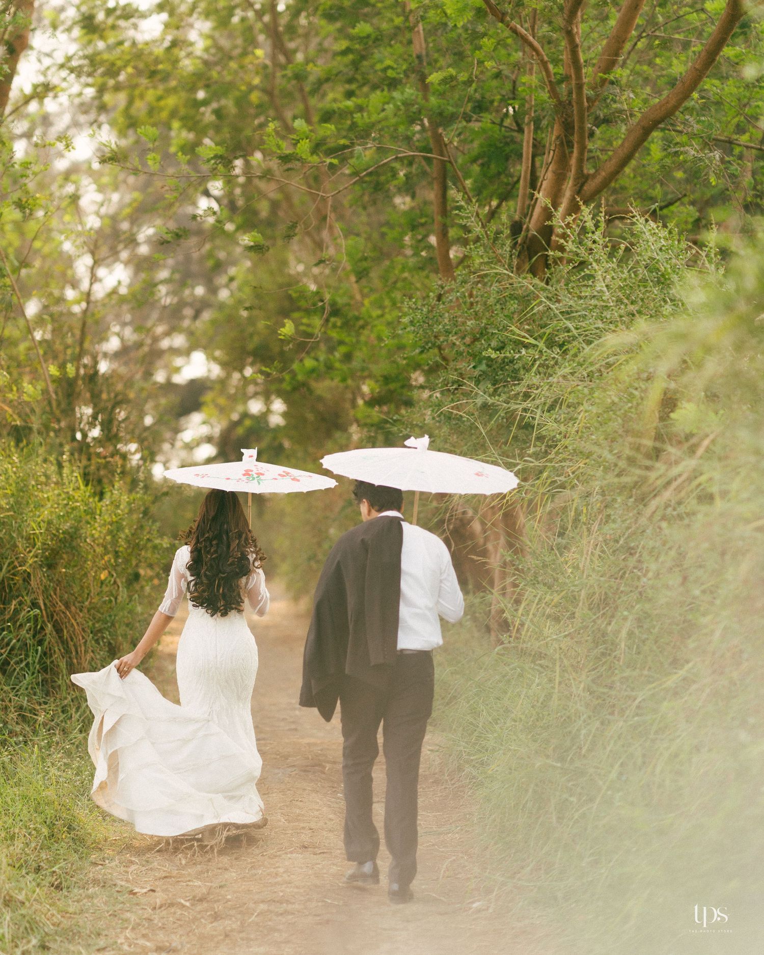 A couple walks down a nature path holding white parasols on their wedding day.