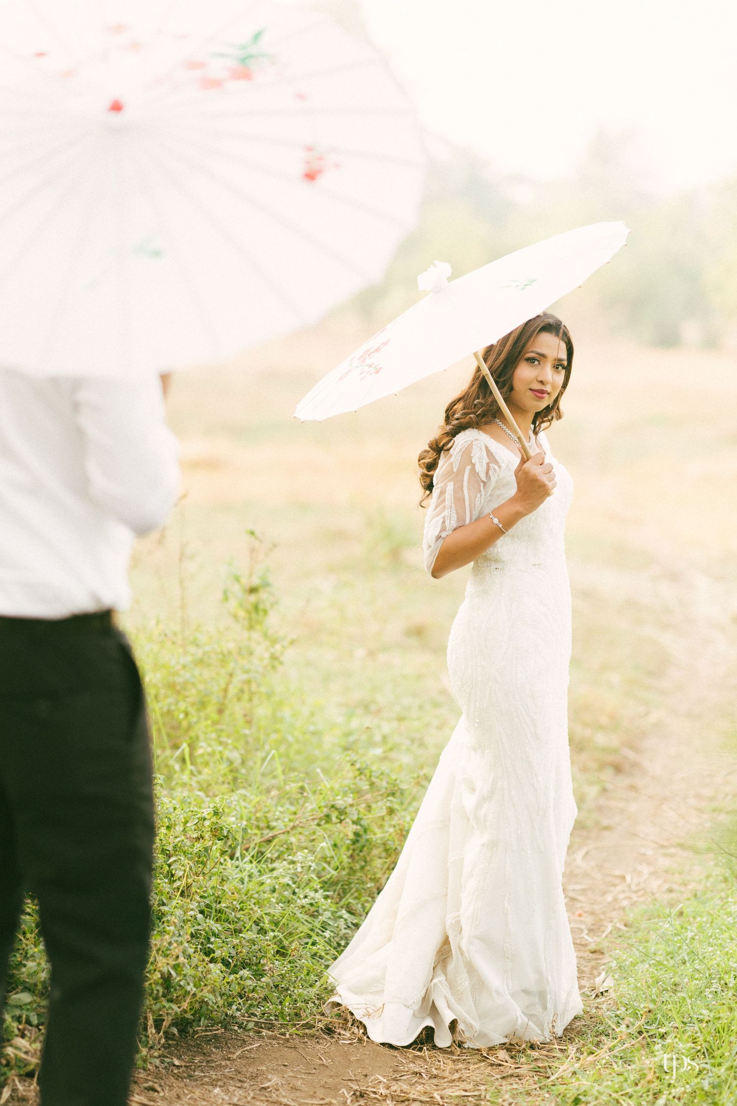 A bride in a flowing white dress poses with a parasol in a sunlit meadow.