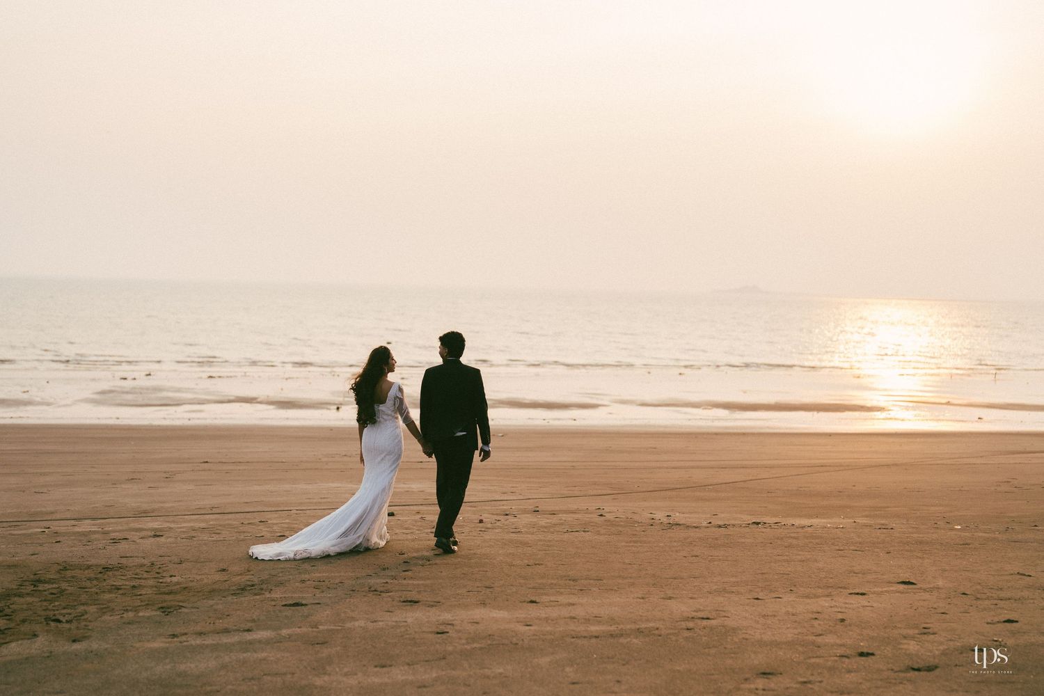 A couple walks along a sunset beach in wedding attire.