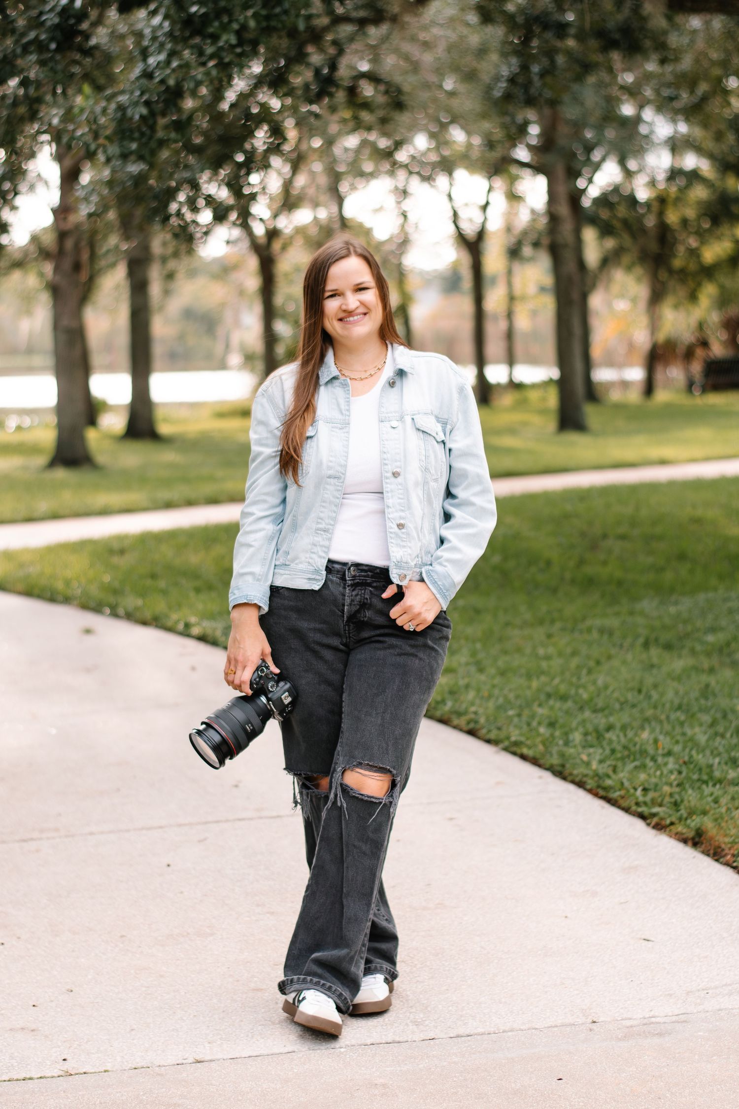 Smiling photographer in light denim jacket holds professional camera on tree-lined path.