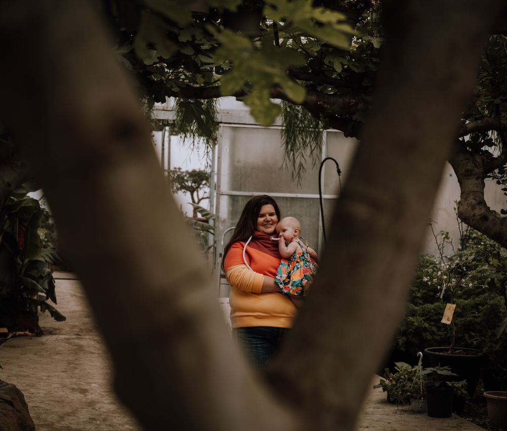 Framed through tree branches, figure in orange sweater holds infant in greenhouse setting.