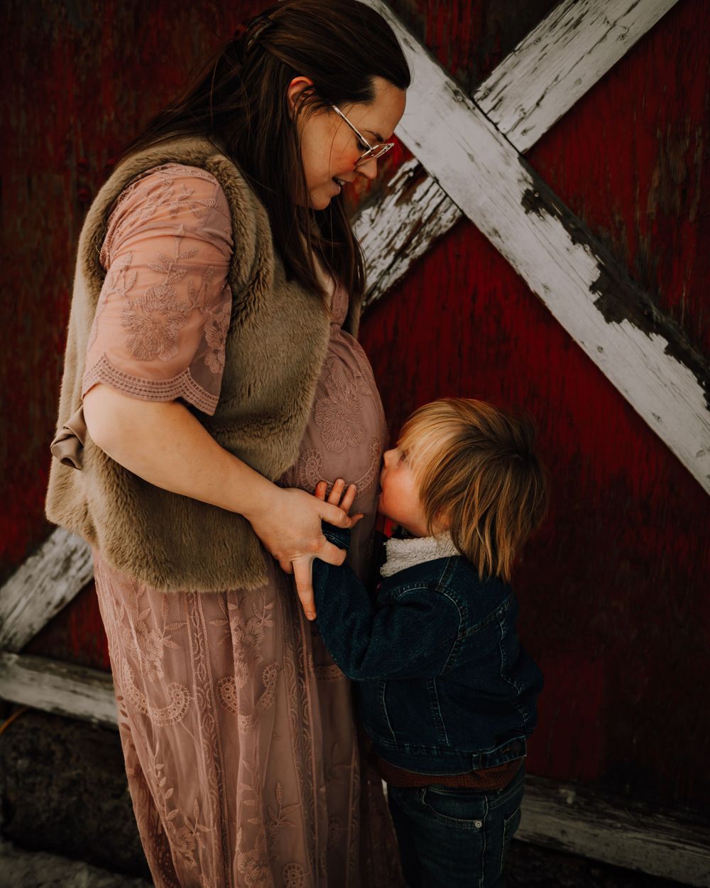 Intimate portrait of parent and child against rustic red barn wall with white cross beam.