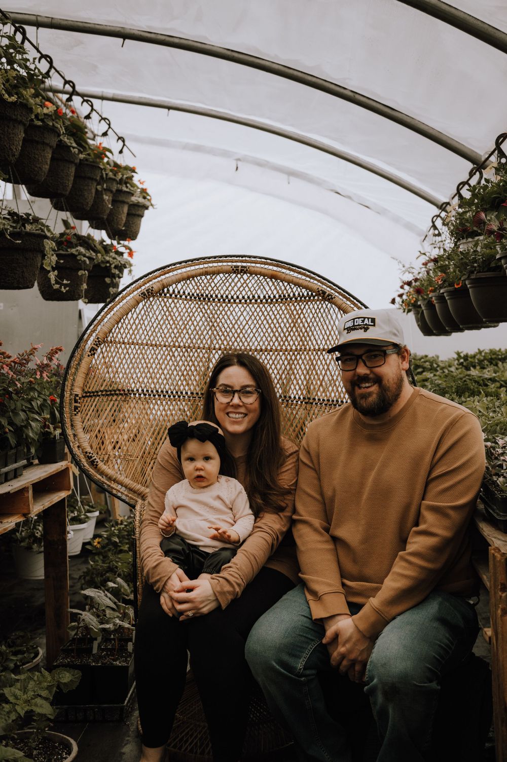 Family sitting together on a woven peacock chair inside a greenhouse with hanging plants.